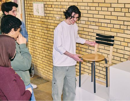 Ein Schüler zeigt vor den anderen Schüler:innen auf sein Workshopergebnis: Eine aus Holz gestaltete mobile Stuhllehne für Hocker